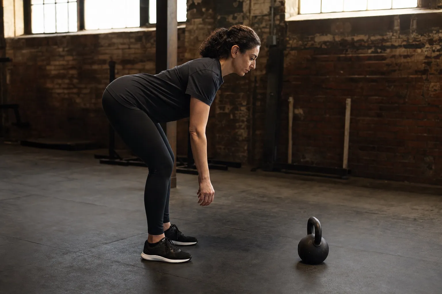 A woman practices an unloaded hip hinge in profile while a kettlebell waits on the floor beside her.